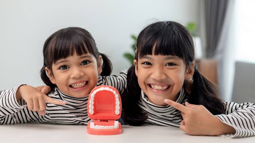 Two young girls are undergoing stage one orthodontics and smiling together while pointing at a small model of teeth.